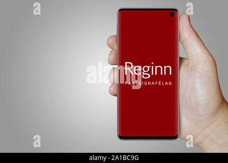 Un uomo possiede uno smartphone visualizzando il logo della società Reginn quotata al Nasdaq Islanda. Sfumatura grigia in background. Credito: PIXDUCE Foto Stock
