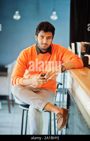 Certi giovani indiani uomo in arancione maglione seduta bancone bar presso il cafe con telefono mobile a portata di mano. Foto Stock