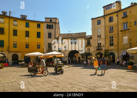 Piazza dell'Anfiteatro, una famosa piazza ellittica a Lucca, con turisti, una famiglia su un cargo bike e un postino su uno scooter, Toscana, Italia Foto Stock