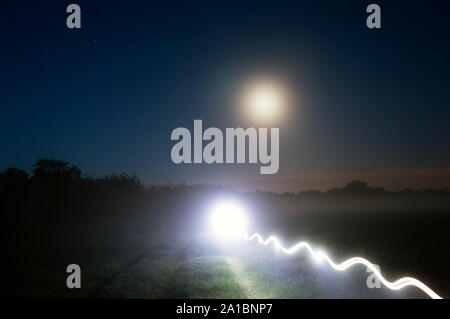 Una brillante luce misteriosa trail su un percorso di paese in una nebbiosa notte di luna brillante nel cielo notturno Foto Stock