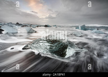 Colpisce il blocco di ghiaccio su una spiaggia nera con surf, tracce di circolazione dell'acqua sulla sabbia scura, in background più blocchi, contrastanti sky con c Foto Stock