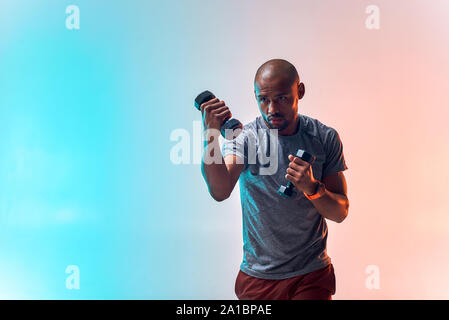 Ottimo allenamento. Muscoloso giovane africano esercizio con manubri in piedi contro lo sfondo colorato. Sport concept. Uno stile di vita sano Foto Stock