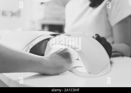 La donna nel salone di bellezza. Mano con manicure fresca nell'UV LED lampada per unghie. Essiccazione del gel sui chiodi, processo closeup. La bellezza e la cura di sé. Tonificazione monocromatica Foto Stock