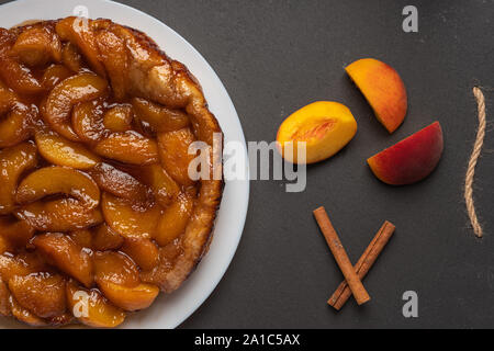 Tarte Tatin con pesche e caramello su una piastra bianca su sfondo scuro. Vicino a fette di pesche e bastoncini di cannella. Vista superiore, orientamento orizzontale Foto Stock