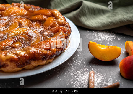 Tarte Tatin con pesche, caramello e zucchero in polvere close-up su una piastra bianca su sfondo scuro con un tovagliolo di lino. Vicino a fette di pesca e Foto Stock