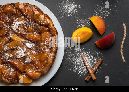 Tarte Tatin con pesche, caramello e zucchero a velo su una piastra bianca su sfondo scuro. Vicino a fette di pesche e bastoncini di cannella. Vista superiore Foto Stock