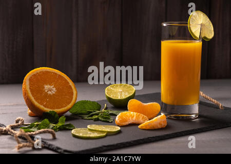 Il succo di arancia in un bicchiere con una fetta di limone su una lavagna nera su legno scuro dello sfondo. Vicino a Orange, menta, fette di mandarino. Cibo sano Foto Stock