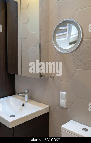 Parte del bagno in una casa privata. Armadietto a parete di piastrelle con specchio. Sotto lavello con rubinetto. Montato a parete round speciale specchio di ingrandimento Foto Stock