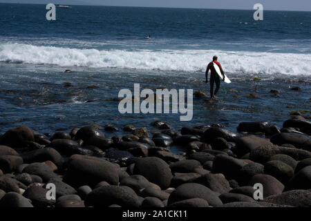 Uomo che cammina tra rocce e carico di acqua la sua tavola da surf per fare surf sulle spiagge di Baja California Messico Foto Stock