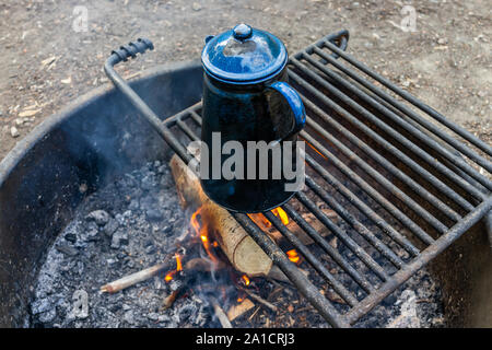 Tè blu bollitore d' acqua sulla griglia in una buca per il fuoco in campeggio con fiamma rosso fuoco che arde in sera Foto Stock