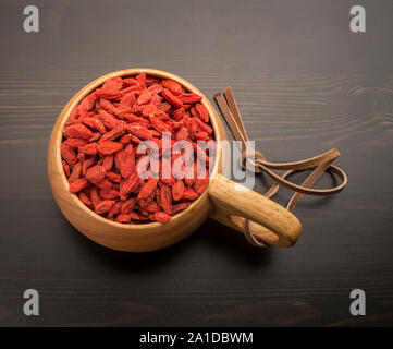 Bacche di Goji in una ciotola di legno con corda in pelle scamosciata, che sorge su una tavola nera Foto Stock