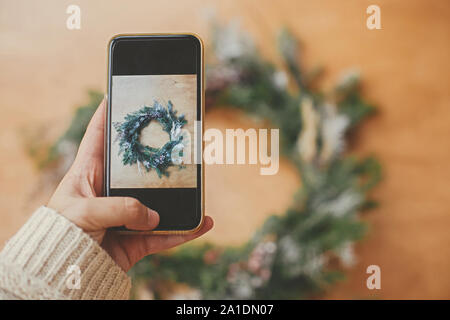 Mano che tiene il telefono e prendendo foto di rustico ghirlanda di Natale lay piatto. Natale mockup. Hipster ragazza facendo foto di Natale corona sullo smartphone Foto Stock