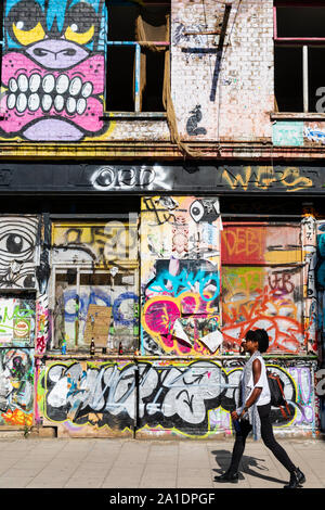Una giovane donna cammina davanti a un edificio abbandonato coperto di graffiti in Hackney Wick, East London, Regno Unito. Foto Stock