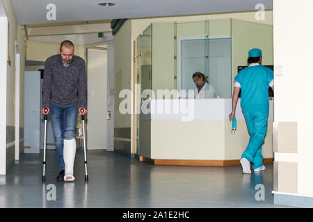 Nel corridoio di un ospedale con un paziente di sesso maschile con gamba rotta, infermiere in background di un medico sulla sua strada Foto Stock