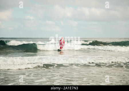 Un giovane uomo surfer vestiti da Babbo Natale sulla spiaggia a fare surf Foto Stock