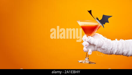 Foto della mummia la mano con il bicchiere di vino e il nero bat vuoto su sfondo arancione in studio. Spazio per il testo, Foto Stock