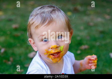 Un adorabile baby boy gode festeggia il suo primo compleanno a una festa con una torta smash di vivacemente colorate torta ghiacciata all'aperto in giardino Foto Stock