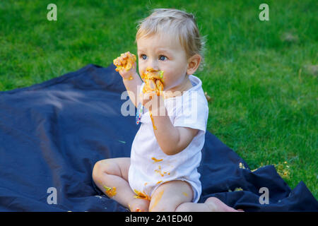 Un adorabile baby boy gode festeggia il suo primo compleanno a una festa con una torta smash di vivacemente colorate torta ghiacciata all'aperto in giardino Foto Stock