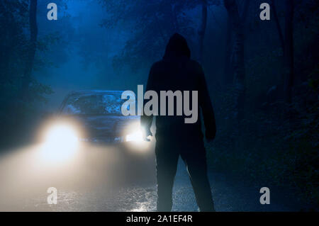 Un lone incappucciati figura in piedi nella parte anteriore dei fari delle auto nel mezzo di una strada di campagna su un losco, inquietanti, moody notte di nebbia Foto Stock