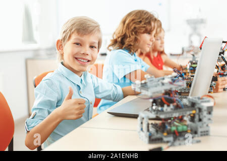 Bambini avente classe di robotica rendendo i robot boy utilizzando laptop guardando la telecamera che mostra il pollice fino a sorridere allegro close-up Foto Stock