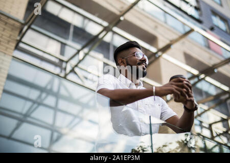 Closeup ritratto di un felice giovane con un drink sul suo balcone, isolata su una città sfondo con strade, edifici e alberi - outdoor Foto Stock