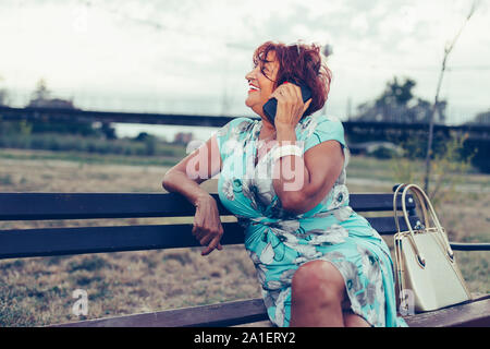 Senior bella donna sta chiamando da un telefono cellulare e parlare mentre è seduto su una panchina Foto Stock