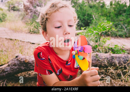 Ragazzo biondo giocando in una foresta con una girandola colorata soffiando aria con faccia buffa. Foto Stock