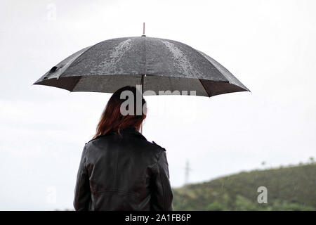 Giovane donna con una giacca di pelle mantiene aperto il nero ombrello in mano sotto la pioggia. Dietro il ragazzo si trova la montagna. Foto Stock