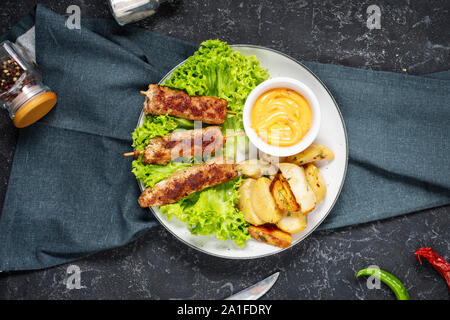 Grigliata di shish kebab servito con patate fritte e salsa di pietra nera dello sfondo. Vista superiore Foto Stock