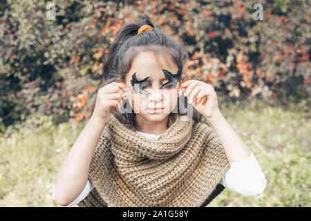 Sorridente bambina che coprono gli occhi con foglie verdi. Kid ritratto d'autunno. Bambino femmina indossando sciarpa lavorata a maglia esterna. Ragazza con felice espressione facciale Foto Stock