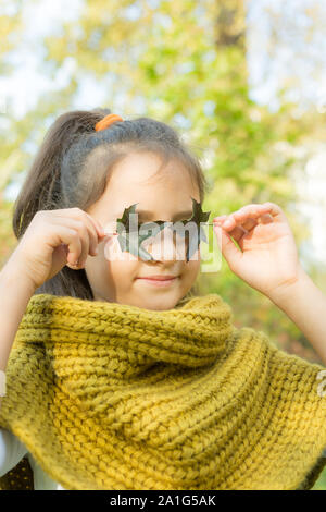 Sorridente bambina che coprono gli occhi con foglie verdi. Kid ritratto d'autunno. Bambino femmina indossando sciarpa lavorata a maglia esterna. Ragazza con felice espressione facciale Foto Stock