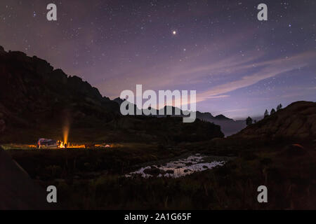 I turisti e i cacciatori sono riscaldati intorno al fuoco di notte nelle montagne sullo sfondo di un cielo stellato e nebbia Foto Stock