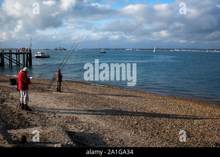 Un uomo che indossa un cappotto rosso sorgeva sulla spiaggia Calshot accanto alla Solent nel canale in inglese mentre un pescatore imposta pronto per giorni di pesca Foto Stock