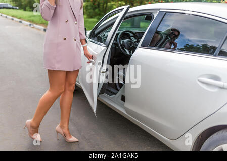 Bella Signora business donna si apre e si chiude la porta auto, in estate in città, business class auto, taxi VIP, rosa tuta, lunghe gambe conciate con tacco alto Foto Stock