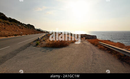 Pomeriggio di sole risplende sulla strada asfaltata che conduce alla spiaggia, il mare in lontananza - tipico paesaggio nella regione Karpas della parte settentrionale di Cipro Foto Stock