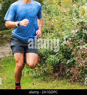 Un runner è su un percorso sterrato con erba in un parco girando intorno a molto alto e cespugli. Foto Stock