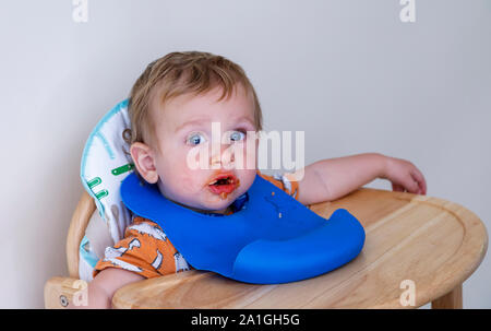 A 1 anni biondo Caucasion baby boy con un confuso faccia rende un pasticcio essendo alimentato nella sua sedia alta con una plastica blu pelican bib come egli impara a mangiare Foto Stock