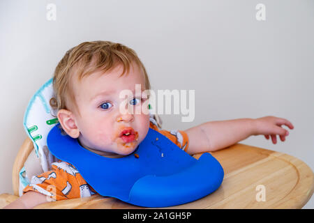 A 1 anni biondo Caucasion baby boy con un confuso faccia rende un pasticcio essendo alimentato nella sua sedia alta con una plastica blu pelican bib come egli impara a mangiare Foto Stock