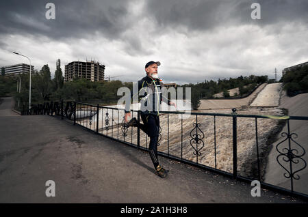 Uomo con barba grigia in costume nero e zainetto in esecuzione sul ponte che attraversa il fiume sporco a sfondo con cielo nuvoloso Foto Stock