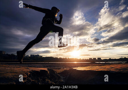 Uomo in silhouette jump correndo contro NUVOLOSO TRAMONTO cielo Foto Stock