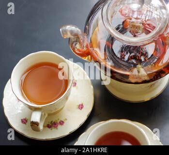 Tè nero con goji (chinese wolfberries) e gelsomino nella teiera in vetro e porcellana tazze con motivo floreale piattini su tavola nera Foto Stock