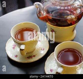 Tè nero con goji (chinese wolfberries) e gelsomino nella teiera in vetro e porcellana tazze con motivo floreale piattini su tavola nera Foto Stock