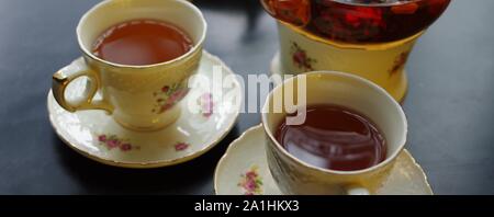 Tè nero con goji (chinese wolfberries) e gelsomino nella teiera in vetro e porcellana tazze con motivo floreale piattini su tavola nera Foto Stock