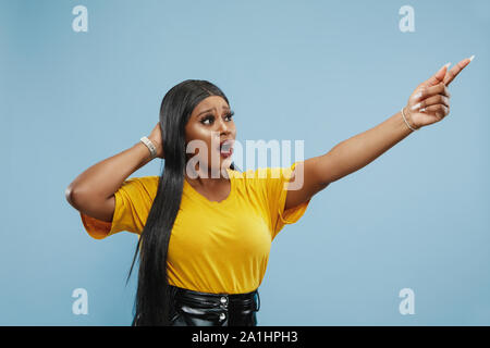 Afro-americano di giovani donna di mezza lunghezza verticale sul blu di sfondo per studio. Bellissimo modello femminile. Concetto di emozioni umane, l'espressione del viso, vendite, annuncio. Puntamento e mostrando qualcosa, sconvolti. Foto Stock