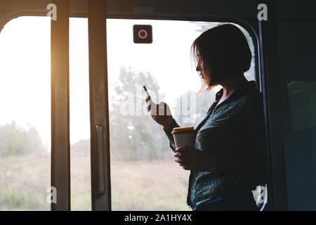 Donna graziosa con un telefono e una tazza di caffè è in piedi in treno Foto Stock