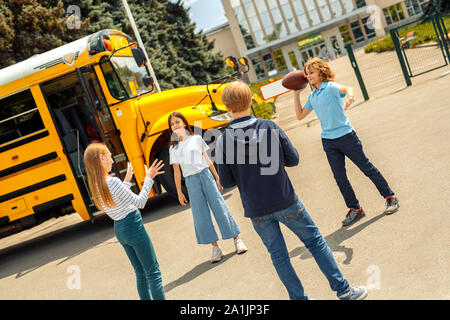 Gruppo di bambini compagni di classe in piedi vicino a scuola bus riproduzione di football americano insieme gettando sfera gioioso sorridente Foto Stock