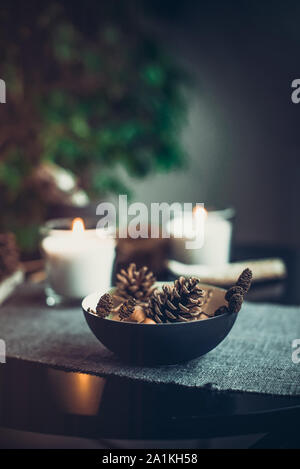 Candele accese e decorazioni naturali di coni, i dadi in vaso nero in tessuto grigio tovagliolo sulla tavola nera. Accogliente atmosfera di casa. Kinfolk Hygge Sl Foto Stock