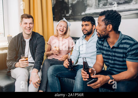 Gruppo di diverse gioiosa amici divertendosi a casa partito, allegramente a ridere, gustando un drink seduti sul divano nel soggiorno. Persone, Tempo libero e ven. Foto Stock