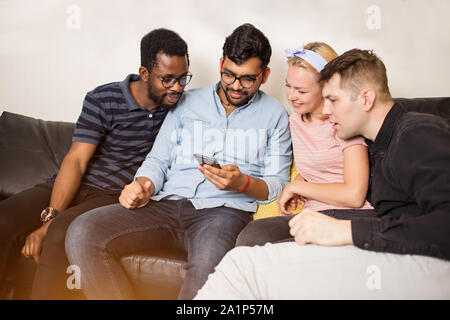 Il gruppo di quattro amici e multirazziale avente il divertimento a casa insieme. Tre uomini e una donna utilizza lo smartphone, guardando un video divertente. Amicizia, il tempo libero e la tec Foto Stock