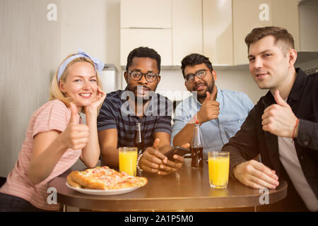 Gruppo di diversi amici seduti a tavola in cucina che mostra il pollice fino gesto, mangiare pizza gustosa e di bere succo di frutta e birra, sorridente guardando la fotocamera. Foto Stock